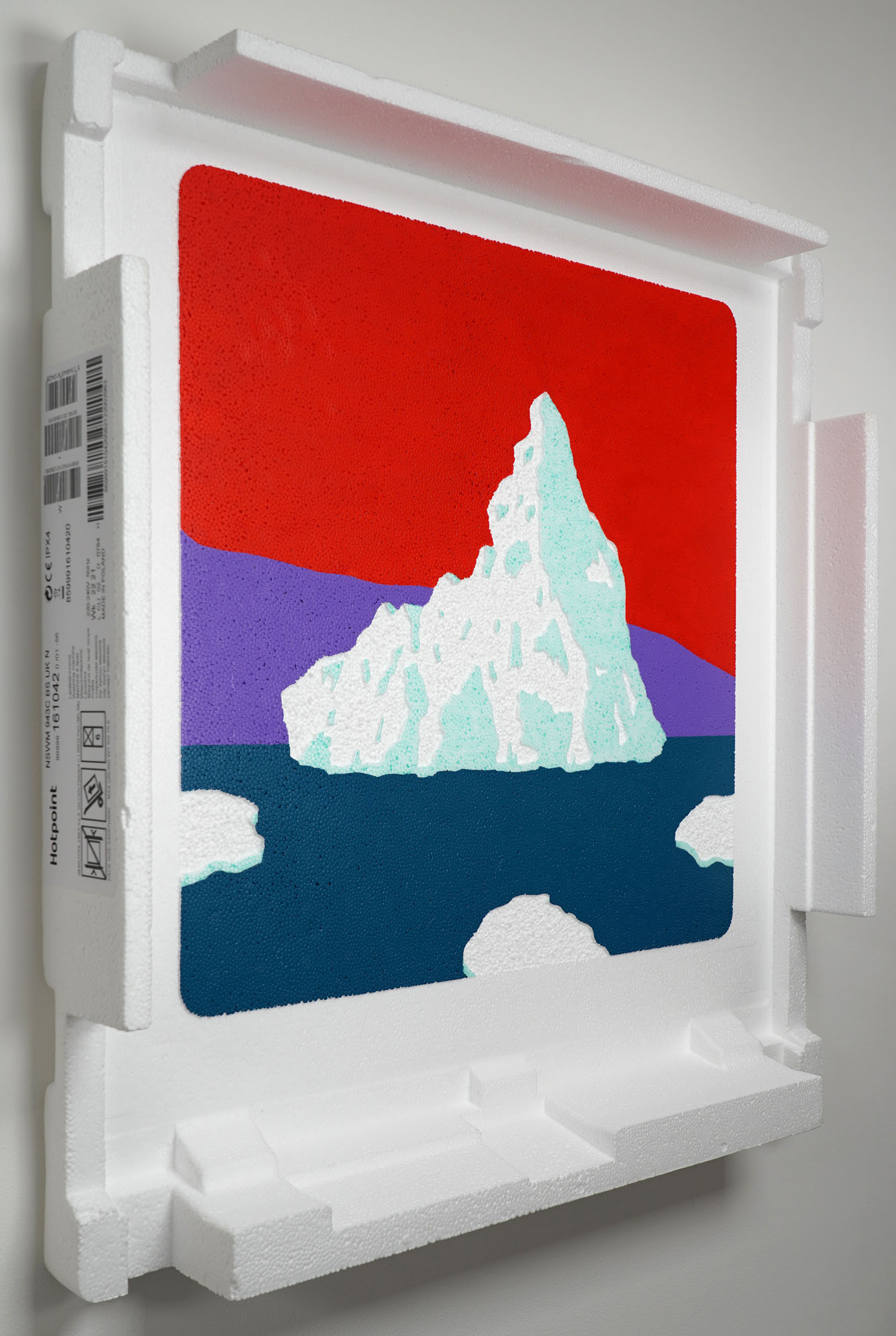 An iceberg with a bright three colour background has been painted and picked onto the surface of some reclaimed polystyrene packaging (with the label 'Hotpoint' on the side).   